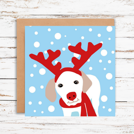 Card with a dog wearing red reindeer antlers and a scarf on a blue snowy background.